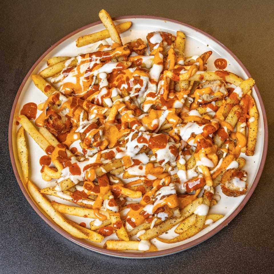 Crispy seasoned fries topped with chicken pieces, drizzled with white and orange sauces on a white plate.