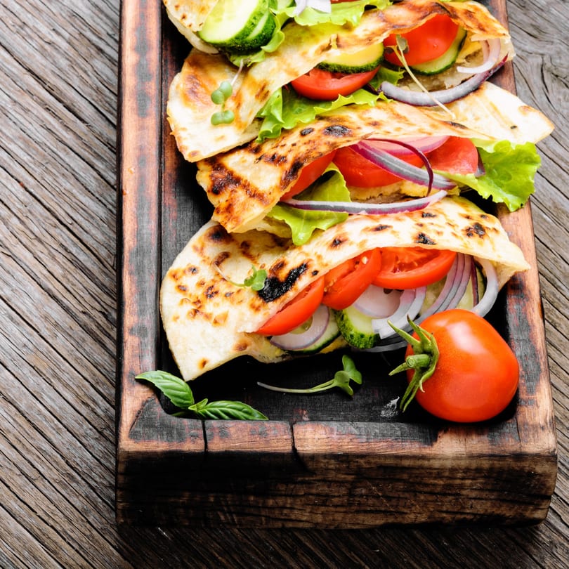 Three grilled flatbread wraps filled with fresh tomatoes, cucumbers, red onions, and lettuce on a wooden board.