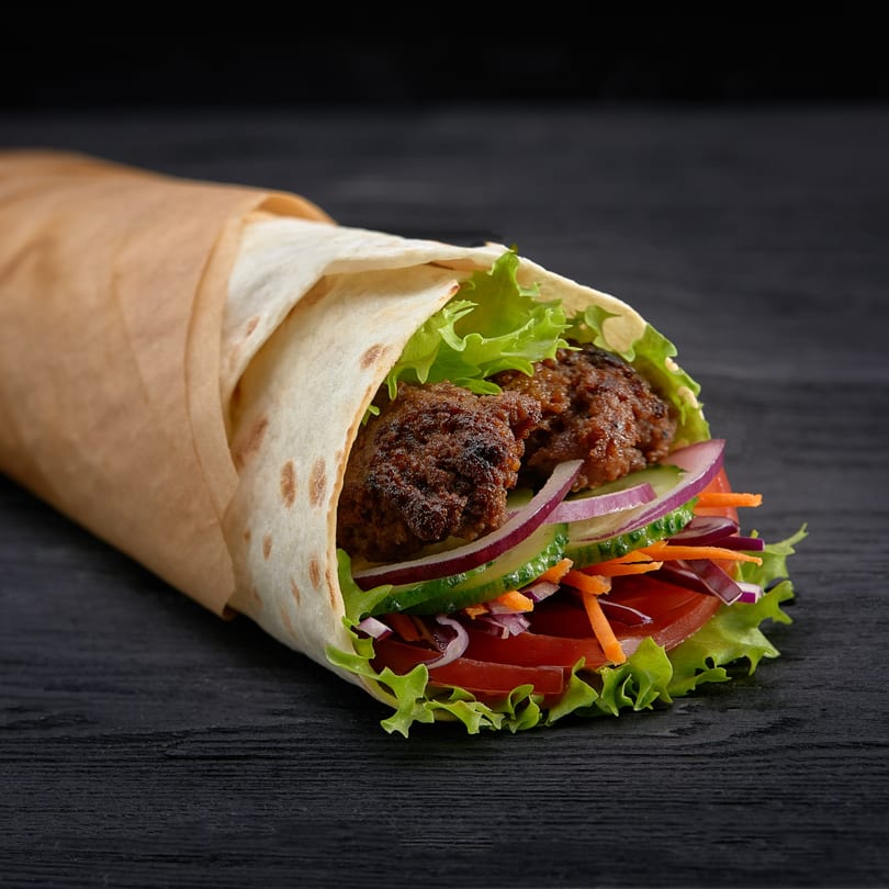 A beef kebab wrap with lettuce, tomatoes, onions, and carrots, partially unwrapped on a dark surface.